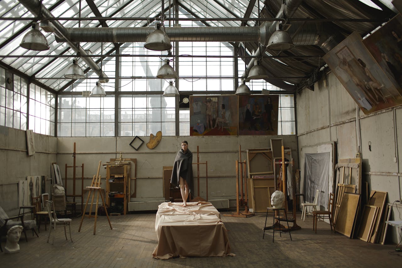 our-story A model stands on a table amidst artistic clutter in a spacious, sunlit studio.